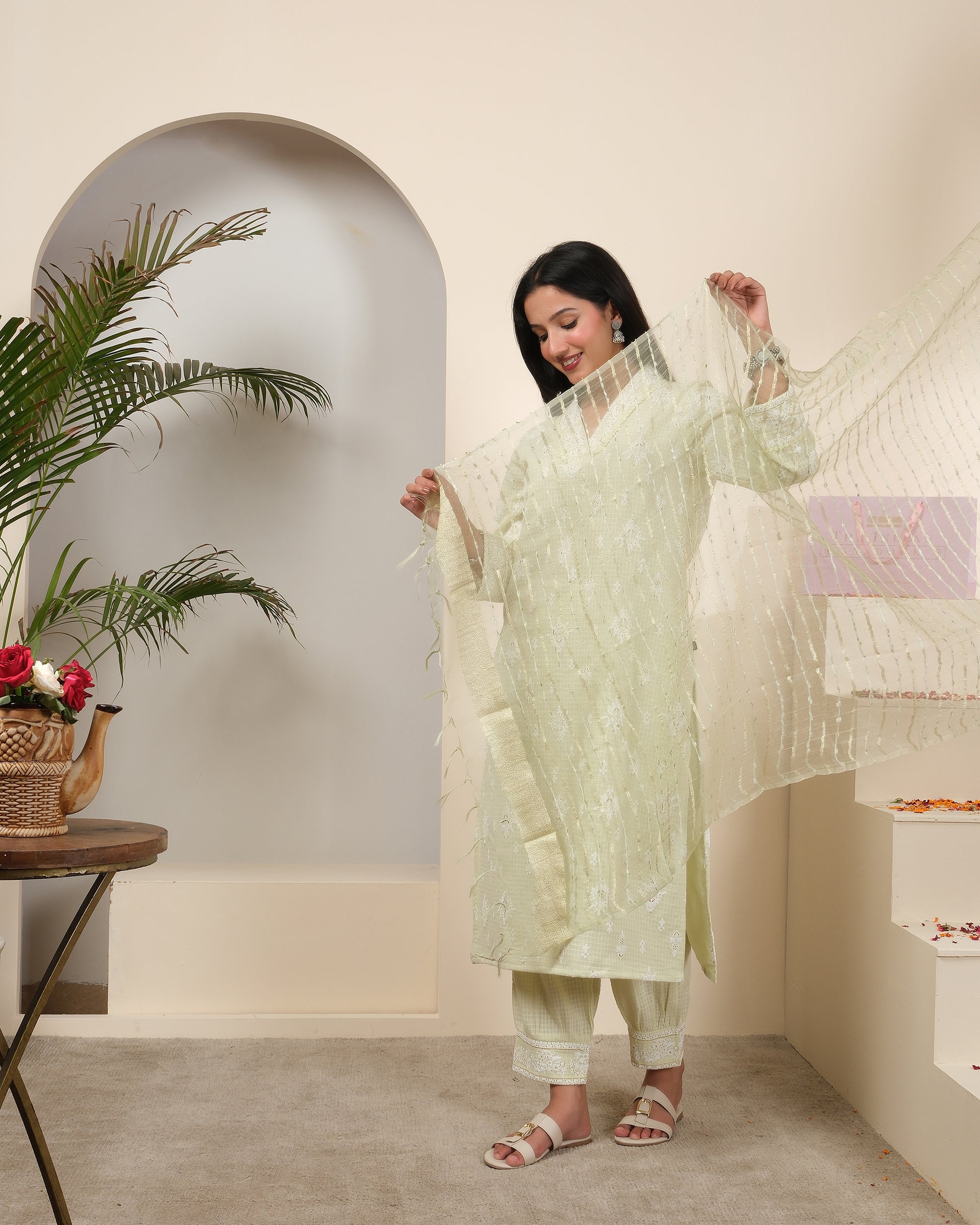 Woman in a light green traditional outfit standing in a minimalistic room with plants and decor.
