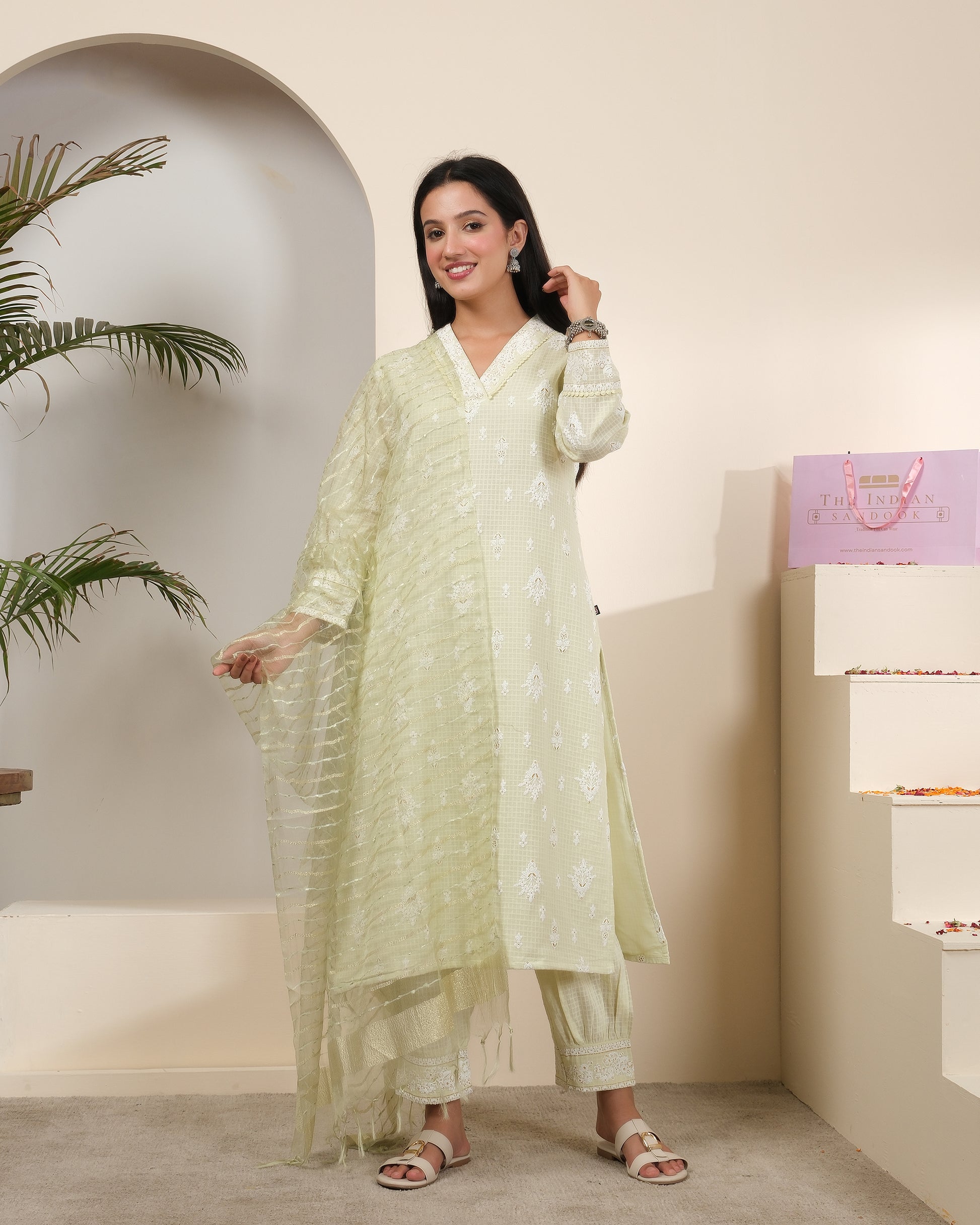 A woman posing in a pastel sage green cotton kurta pant set with a sheer, shimmering striped dupatta featuring tassels